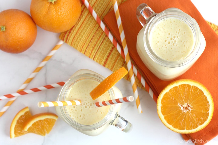 Two smoothies served with festive straws, surrounded by oranges and by orange and pineapple colored cloth.