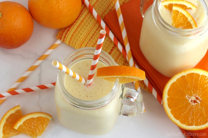 Overhead of two smoothies in glass mugs with orange striped paper straws and extra oranges laying around.