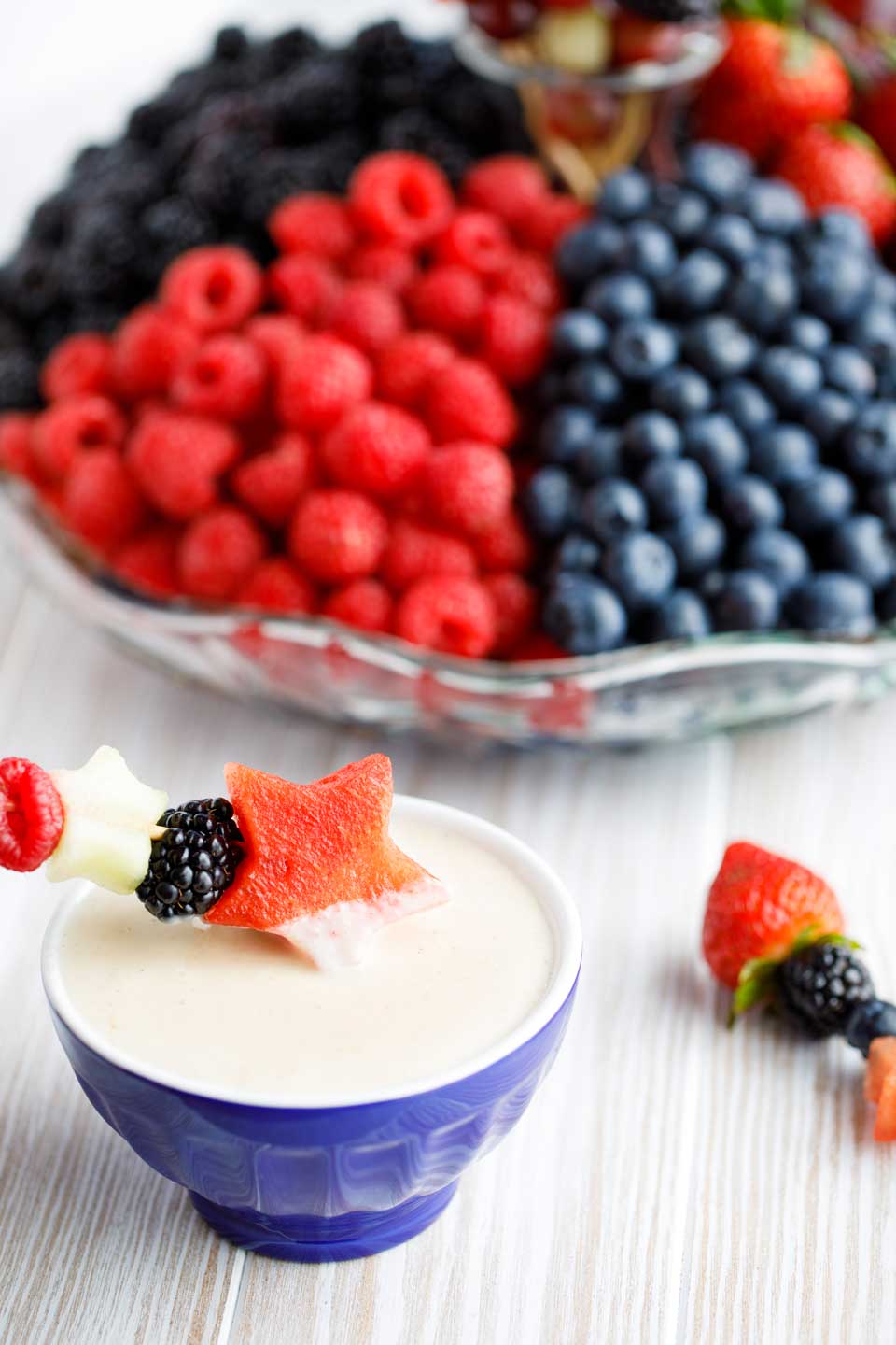 fruit dip in a blue bowl with a red, white and blue -themed fruit kabob dipping into it and a fruit tray of red and blue fruits in the background