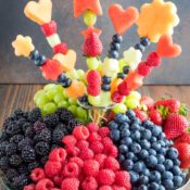 A heaping fruit tray with piles of blackberries, raspberries, blueberries, strawberries and green grapes, and a fruit bouquet at its center
