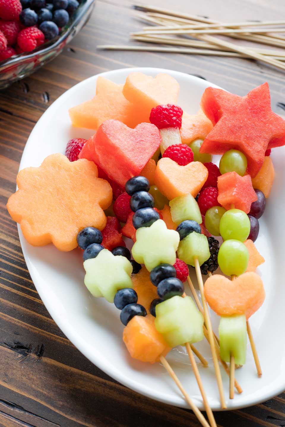 colorful fruit skewers piled on an oval, white platter with additional fruit and empty skewers in the background