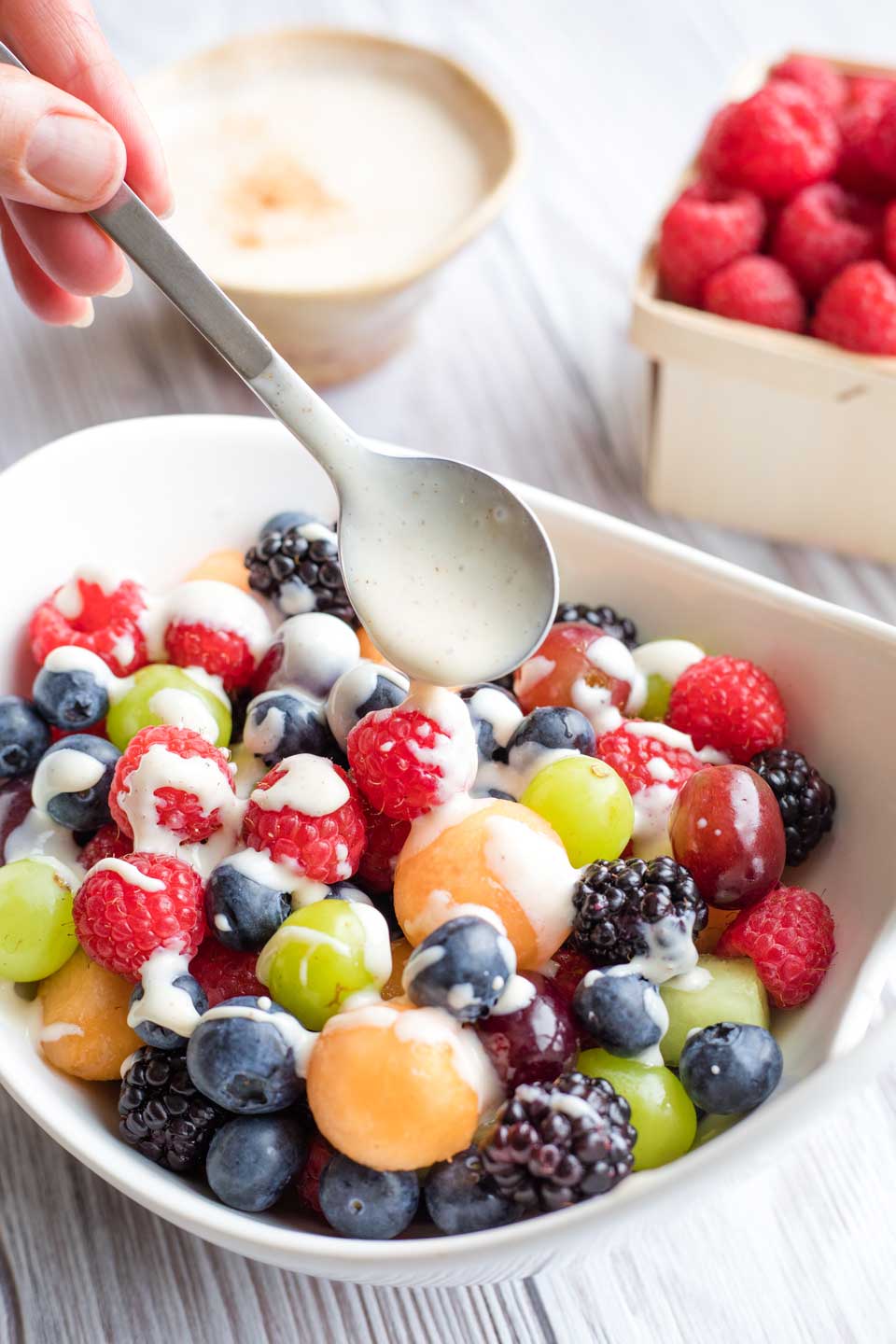 a white bowl filled with colorful fruit salad, with a hand holding a spoon that's drizzling this fruit dip over top of the salad like a dressing