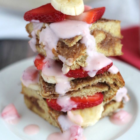 side view of three pieces of this french toast casserole, stacked on top of each other on a white plate, layered with fresh strawberries and bananas and drizzled with fresh strawberry topping sauce