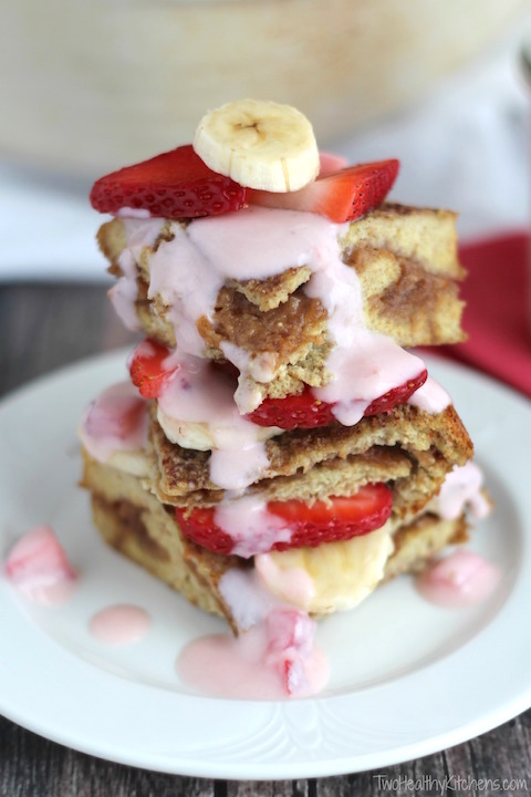 side view of three pieces of this french toast casserole, stacked on top of each other on a white plate, layered with fresh strawberries and bananas and drizzled with fresh strawberry topping sauce
