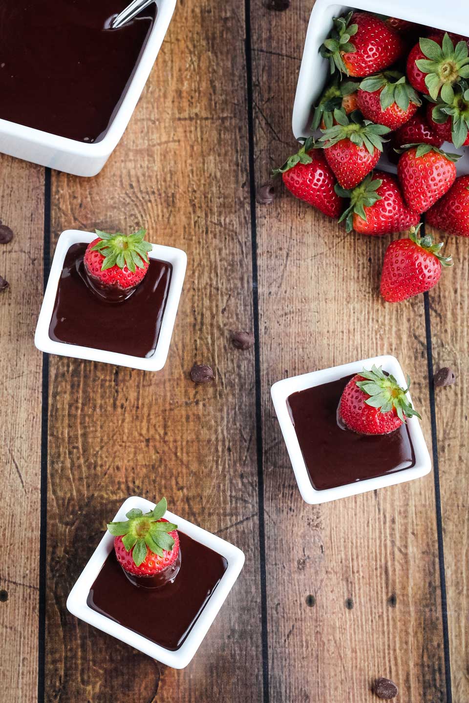 overhead of 3 square bowls of dip with a strawberry dunked in each; a larger serving bowl of Chocolate Dip in the background, and also a cascading container of strawberries