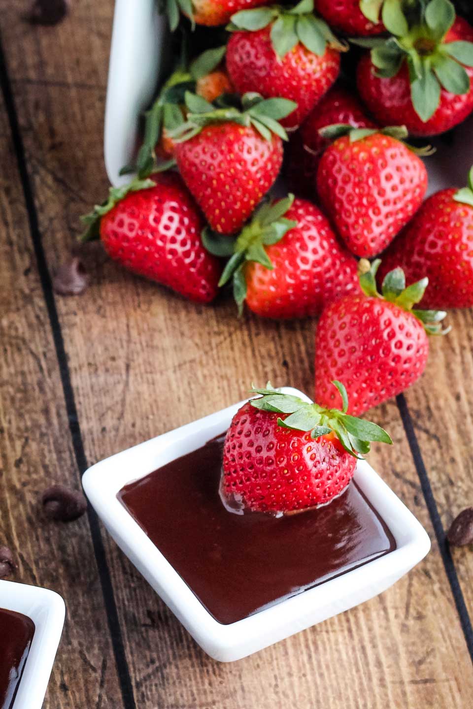 container of strawberries tipped over and cascading across the background, with a small square dish of Chocolate in the foreground, with a strawberry in it