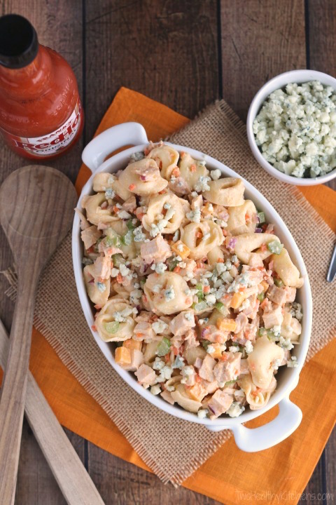 overhead of bowl filled with pasta salad, surrounded by small bowl of blue cheese, serving utensils, and bottle of hot sauce