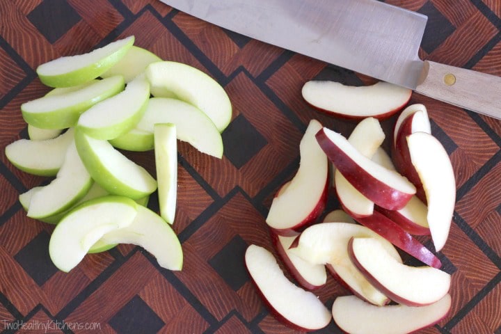 One pile of sliced green apples alongside a pile of slice red ones on a cutting board with knife.