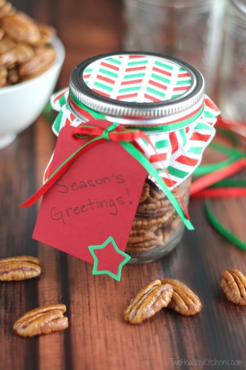Candied nuts packaged as Christmas gift in in glass jar with tag "Season's Greetings!" and extra pecans nearby.