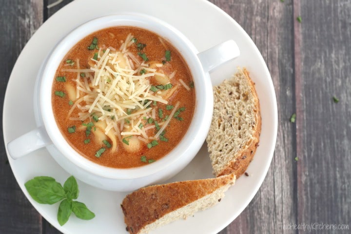 Flatlay of tomato soup in white handled crock on plate with bread wedges and basil sprig.