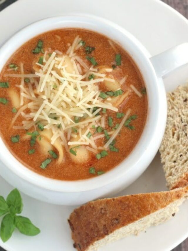 Close crop overhead of tomato soup in white pottery bowl with bread and basil alongside.