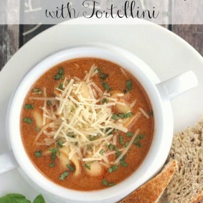 Soup in white crock on plate with bread and basil and text overlay "Tomato Basil Soup with Tortellini".