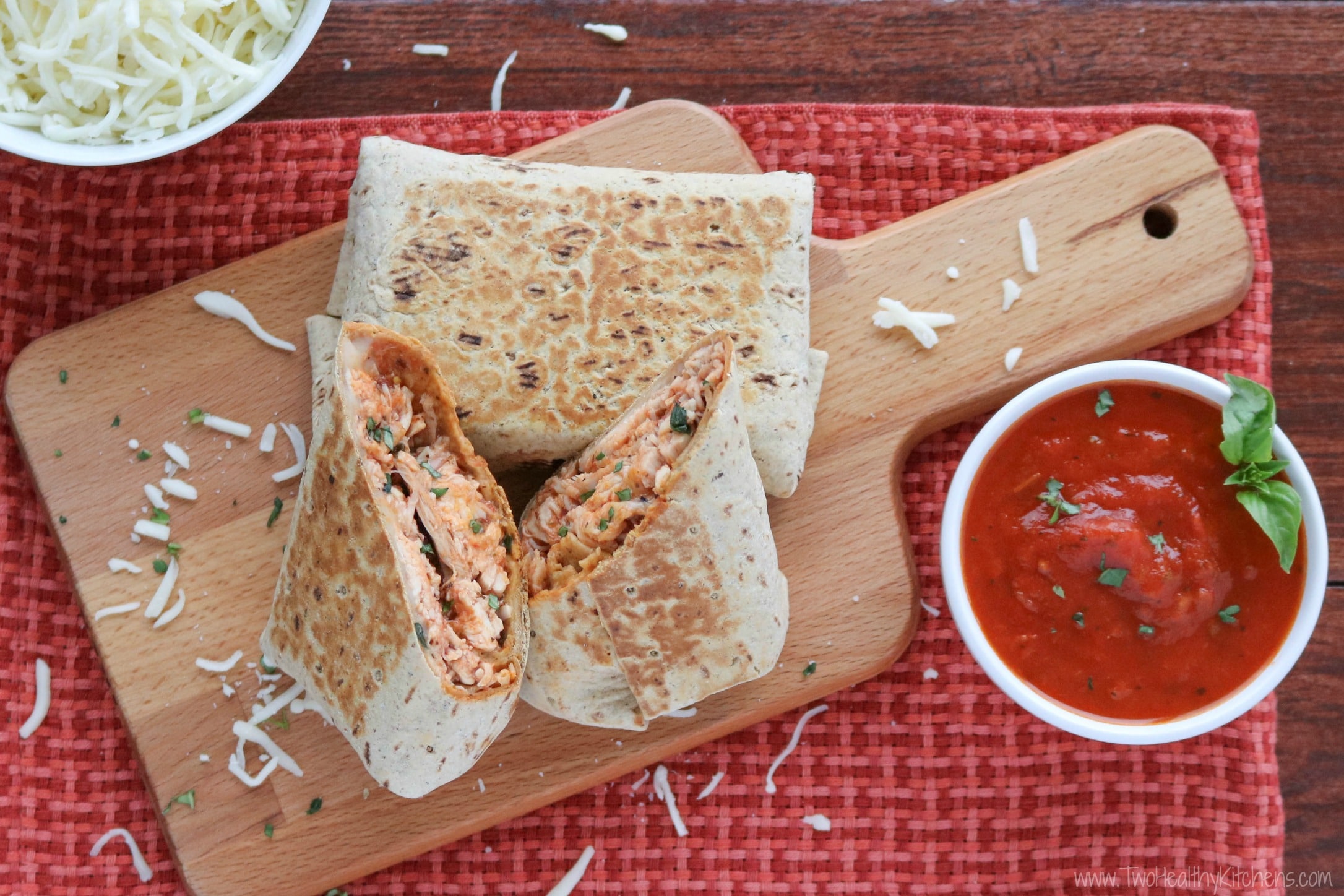 Two cooked wraps on wooden board, one whole and one cut open upright so you can see the chicken parmesan filling, with marinara and extra cheese alongside.