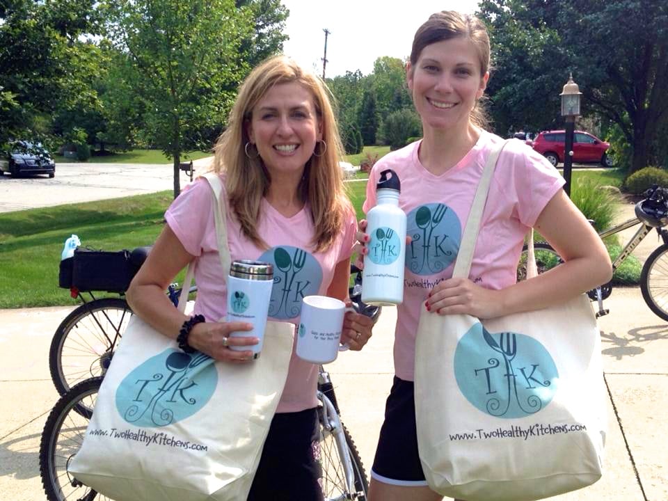 Shelley (L) and Gretchen (R) in pink Two Healthy Kitchen logo shirts, holding bags and various travel mugs with THK logos.