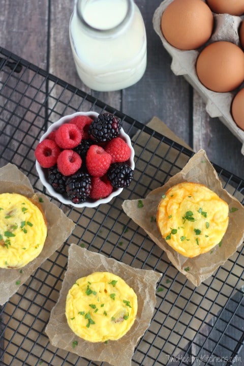 Overhead of 3 egg cups on squares of parchment on baking rack; bowl of berries, bottle of milk and fresh brown eggs nearby.