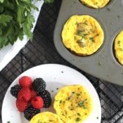 Flatlay scene with 2 egg cups and berries on small white plate on baking rack; muffin tin with more egg ups and fresh parsley surrounding.