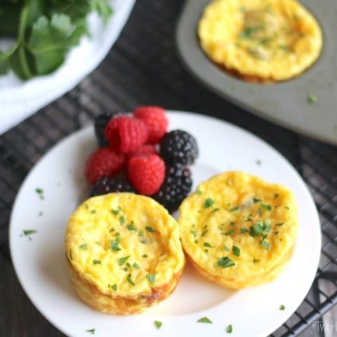 Two egg cups on small white plate with berries, muffin tin of more egg cups in background.