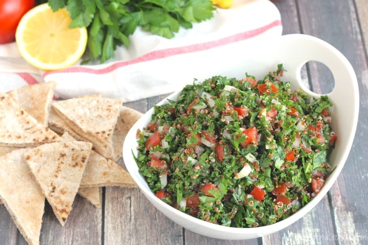 Tabouli piled in curved white serving bowl next to pita triangles; extra ingredients in background.