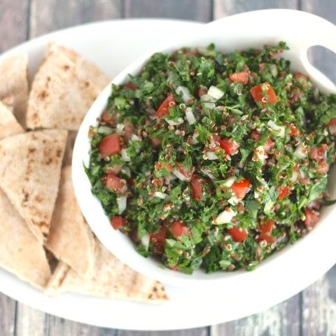 Curved serving bowl of quinoa tabouli on white platter surrounded by pita wedges.