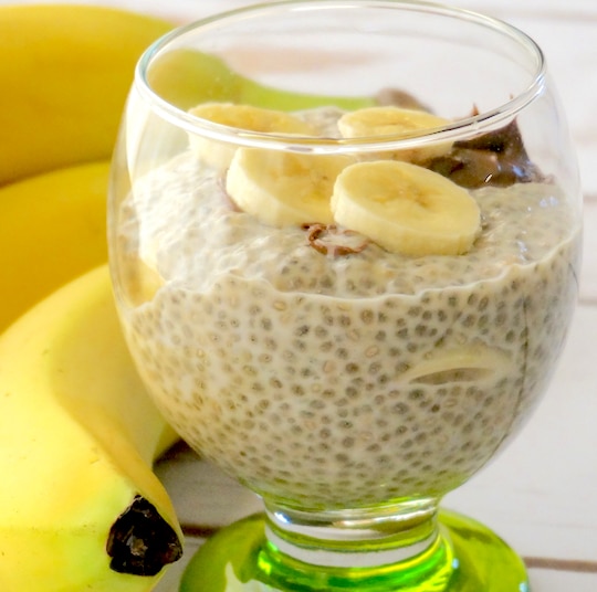Breakfast pudding in green-stemmed parfait glass with whole, unpeeled bananas behind.