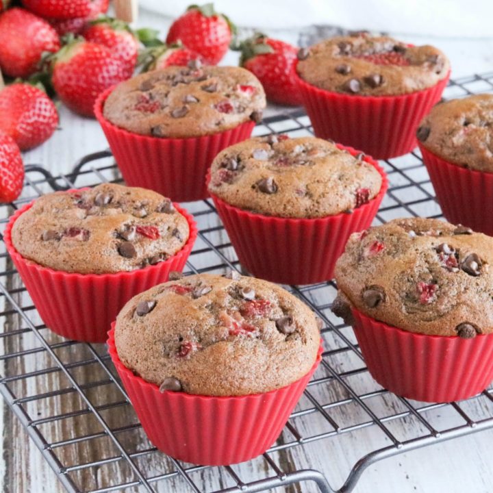 Eight muffins in red silicone liners on cooling racks with fresh strawberries cascading out of pint basket in background.