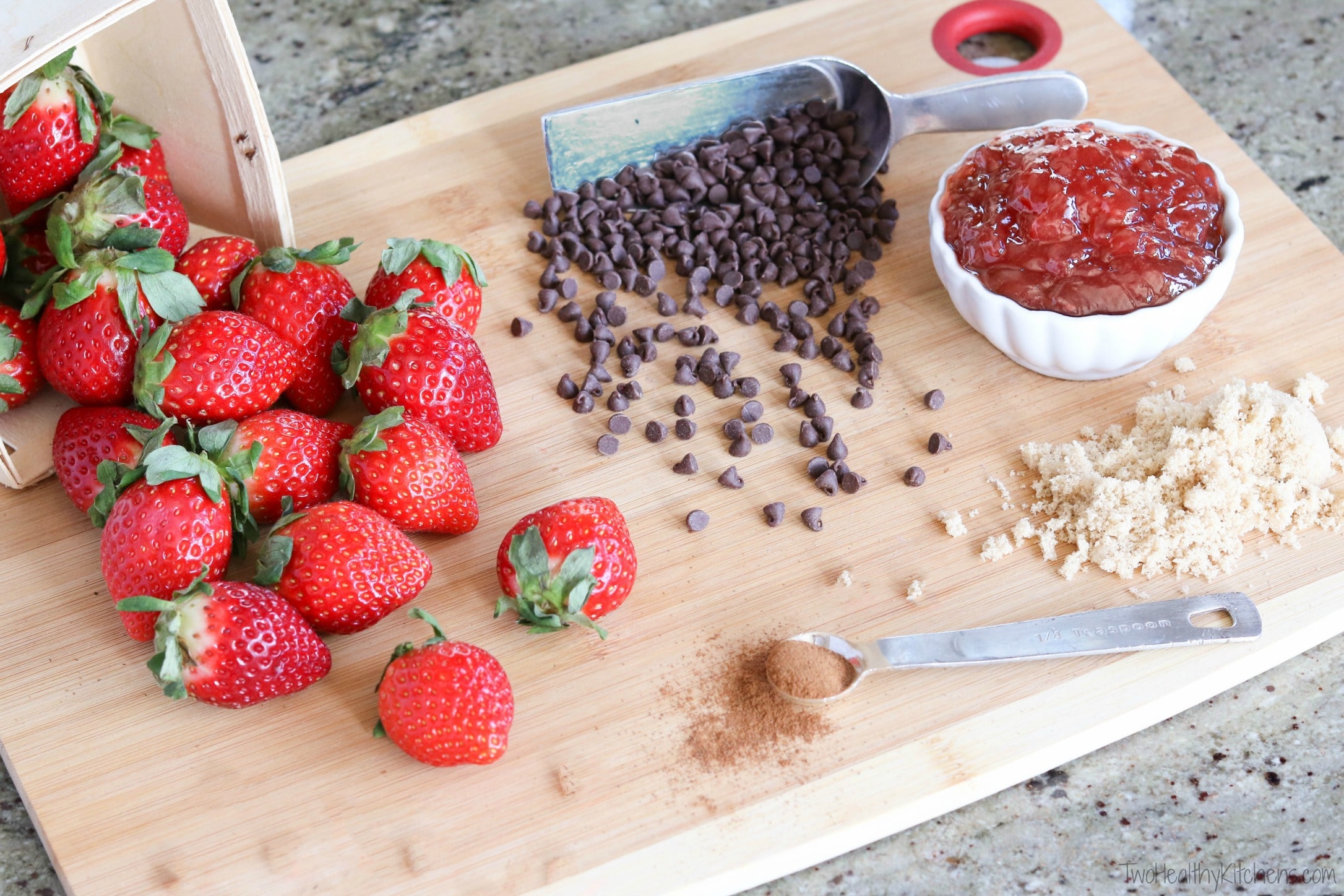 Chocolate chips spilling out of scoop, fresh strawberries cascading out of basket, bowl of strawberry jelly, measuring spoon of cinnamon, and small pile of brown sugar on bamboo cutting board.
