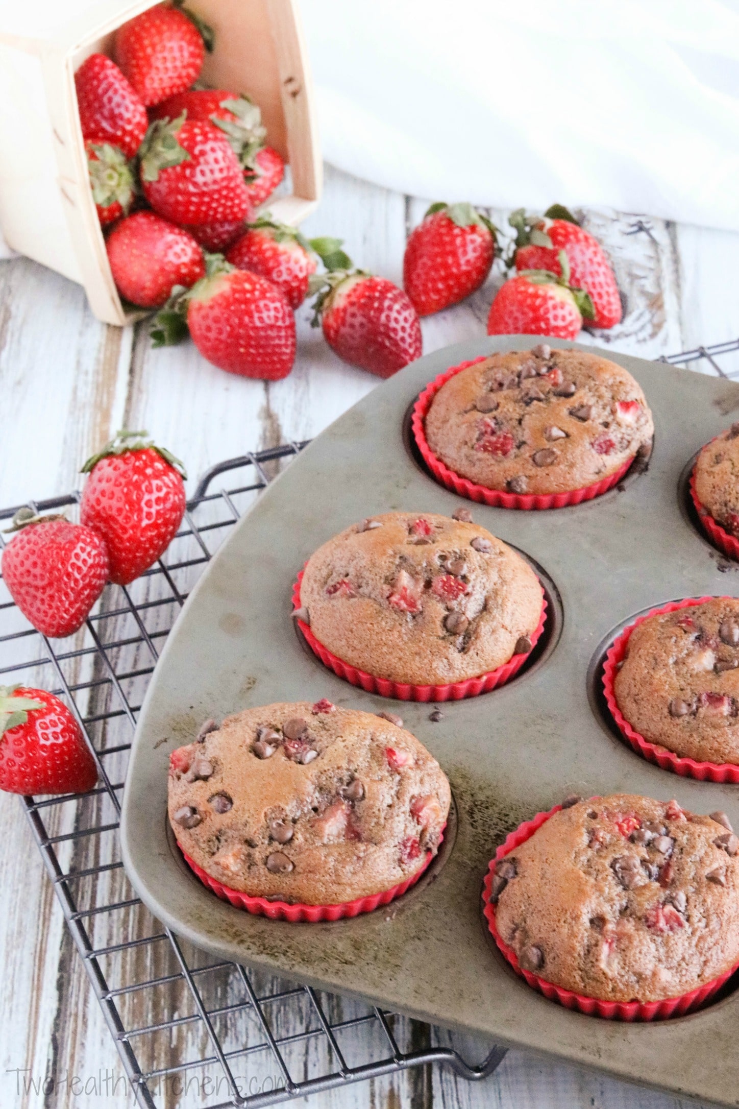 Left side of muffin tin with baked chocolate muffins still in, laying diagonally across cooling rack with strawberries in background.