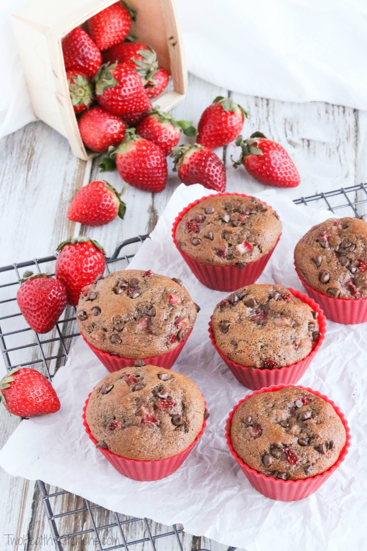 Six of the muffins in red liners on a cooling rack lined with white parchment, with fresh strawberries cascading nearby.
