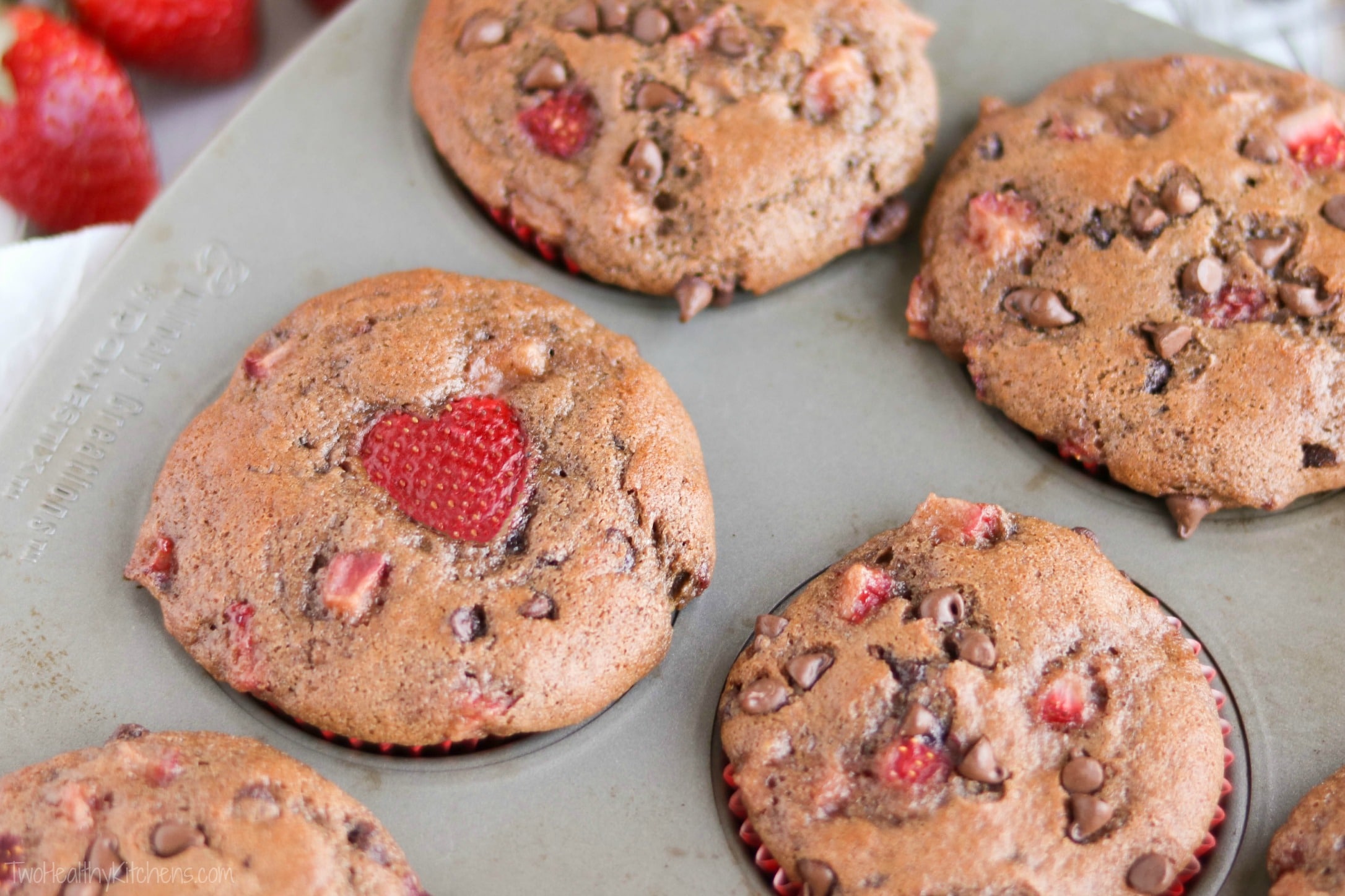 Closeup of baked muffins still in tin, focused on one with a heart-shaped strawberry amidst the chocolate chip topping.