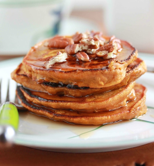 Stack of 4 pancakes on plate with green-handled fork, topped with syrup and nuts.