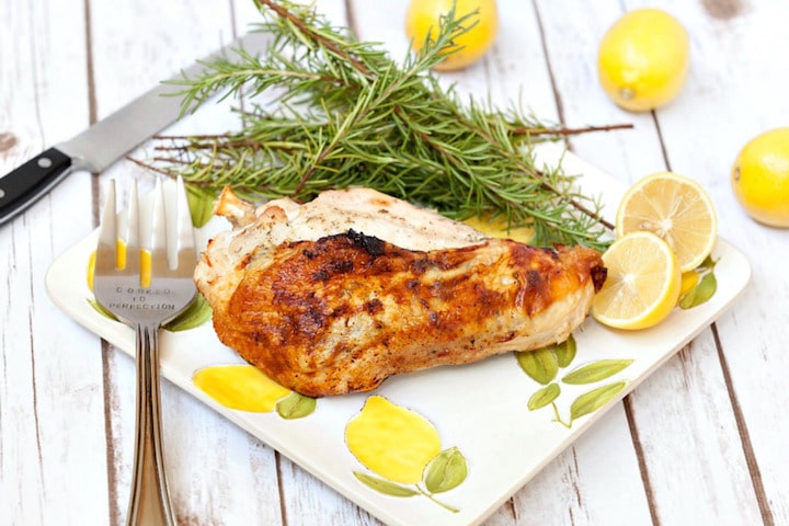 Cooked chicken breast on square, lemon-printed plate with several rosemary sprigs and halved lemon.