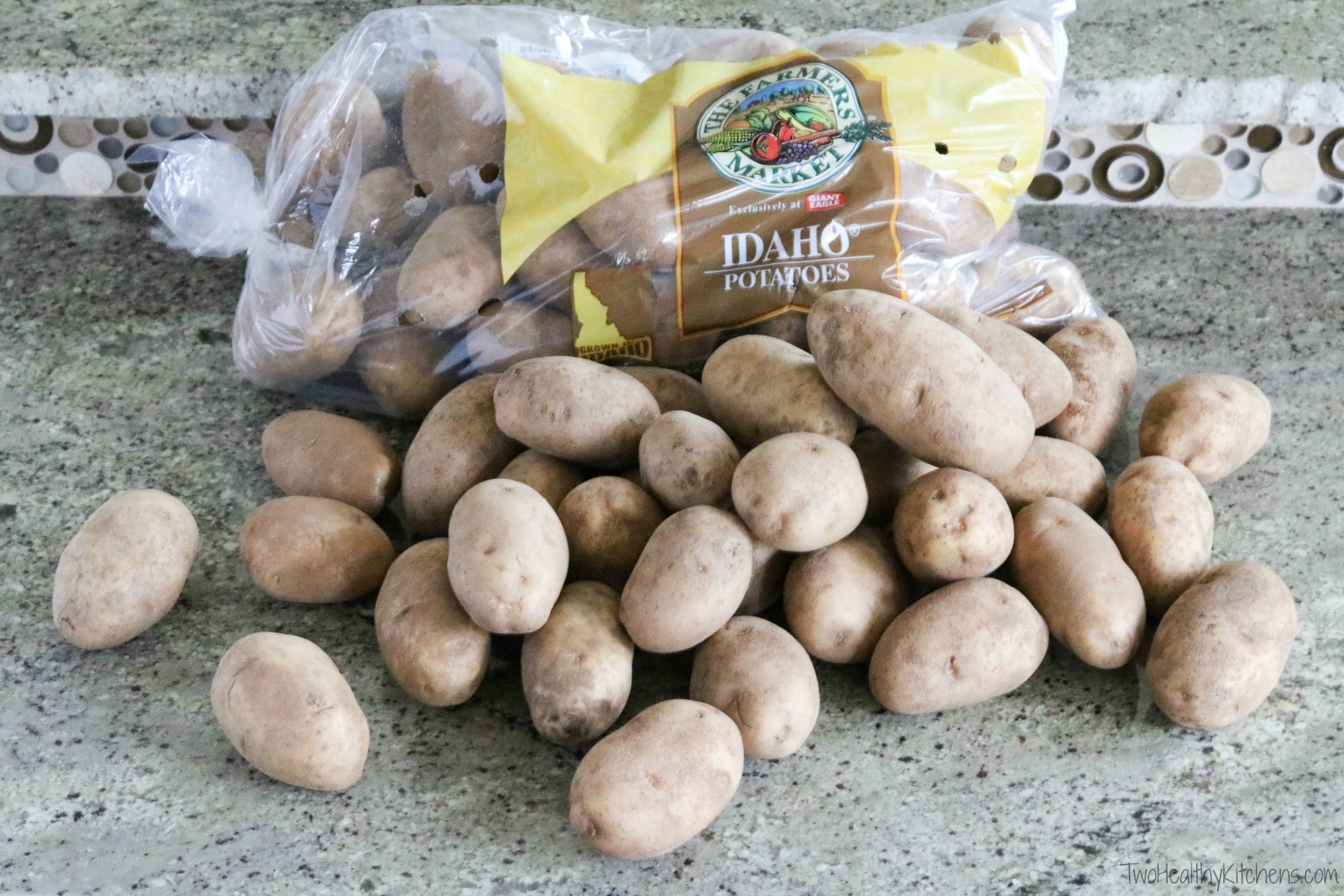 Big bag of russet potatoes on kitchen counter with huge pile of loose potatoes stacked in front of it.