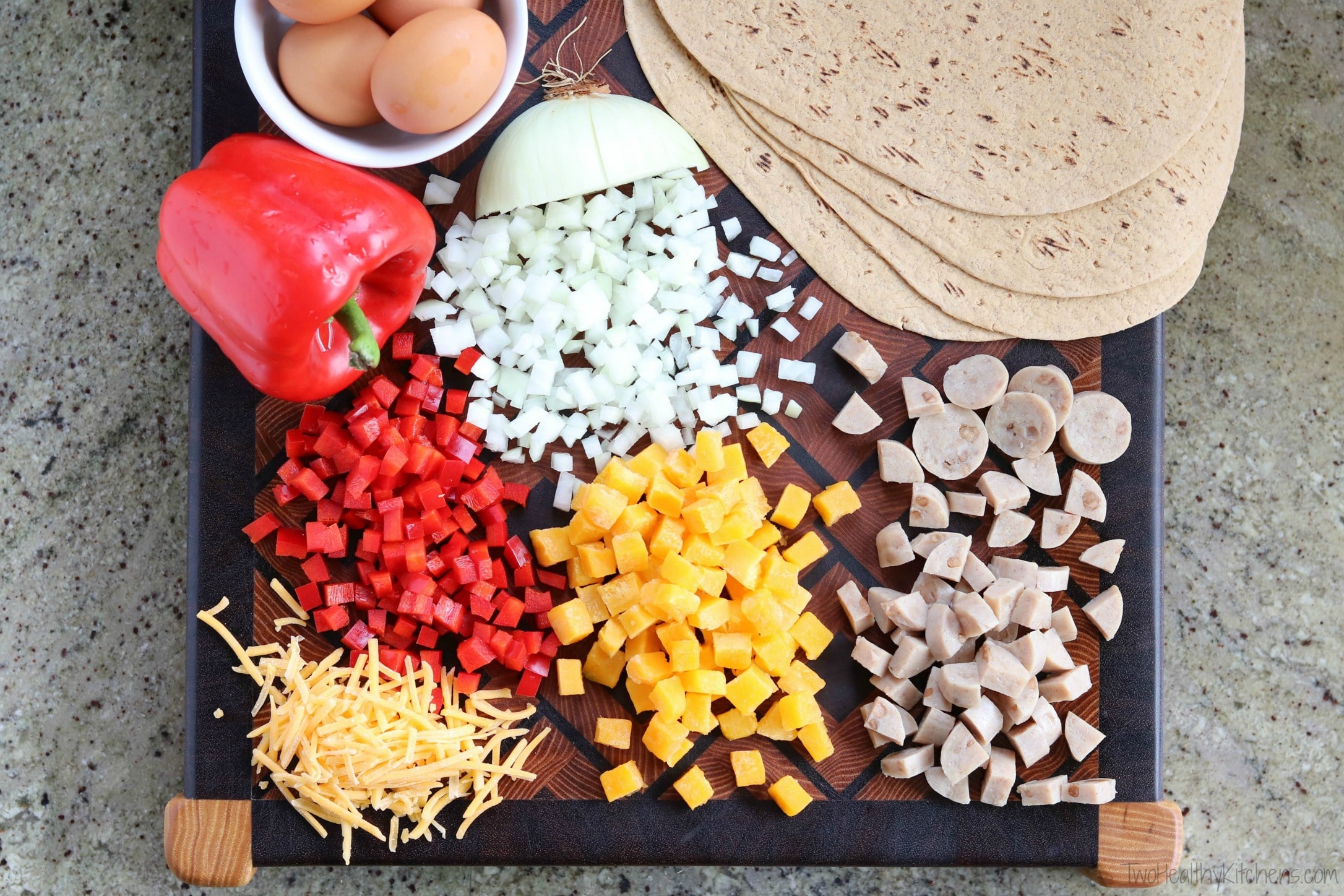 Burrito ingredients chopped in piles on cutting board, with whole red pepper, bowl of brown eggs and wraps.