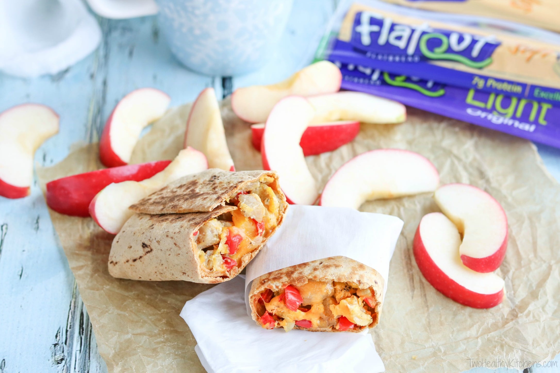 Burrito cut in half on parchment with apple slices, coffee mug and packages of Flatout wraps nearby.