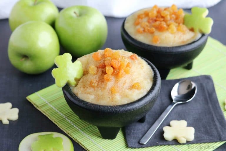 Two black bowls of applesauce on green placemat with black napkin, spoon, and green apples surrounding.