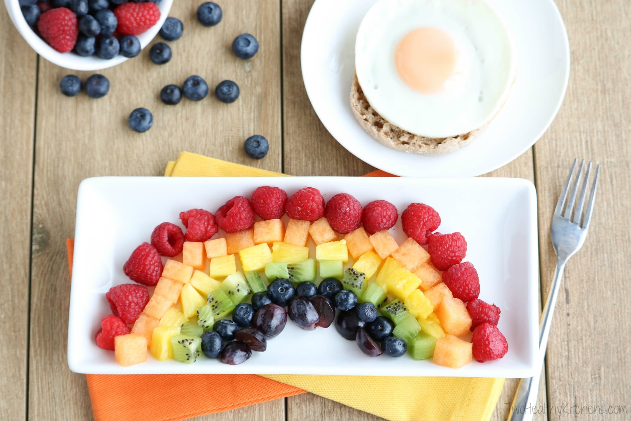 Small white rectangular plate with fruit rainbow, with egg-topped English muffin on round plate behind, plus fork and bowl of berries.