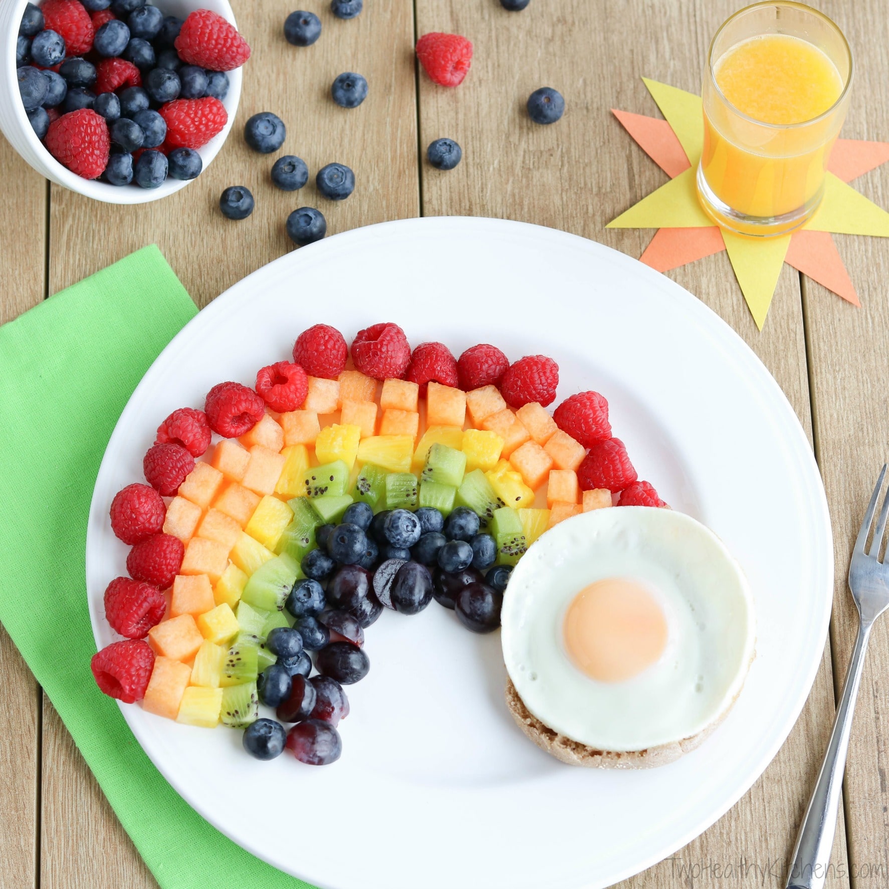Fruit rainbow on white plate with egg-topped English muffin, green napkin, fork, OJ on sunshine paper and bowl of berries.