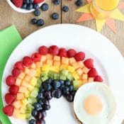 Overhead of fruit rainbow with egg on white plate, with glass of orange juice, green napkin and extra berries.