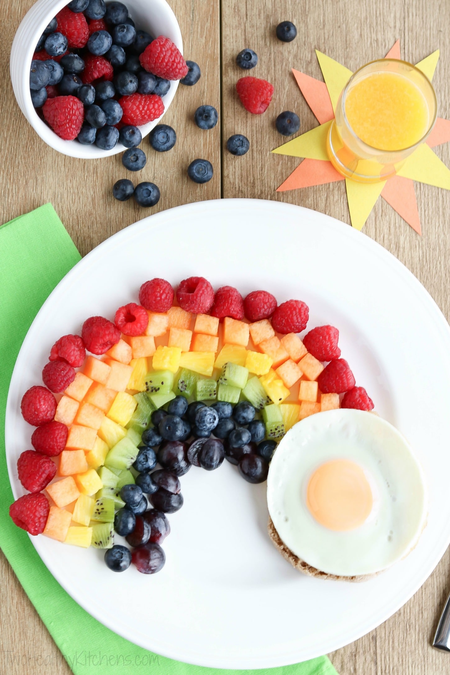 Overhead of fruit rainbow with egg on white plate, with glass of orange juice, green napkin and extra berries.