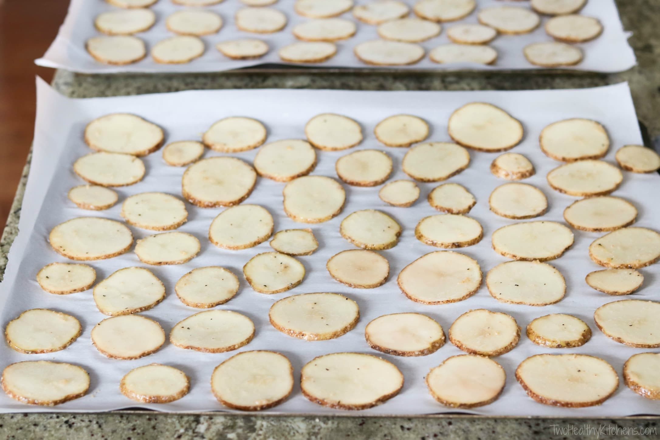Two parchment-lined sheet pans with unbaked potato slices spread out.