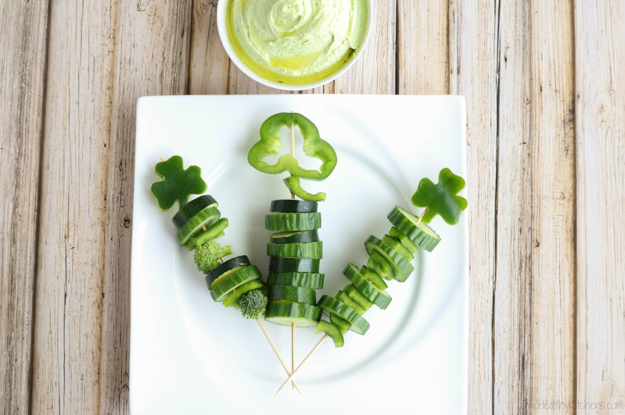 3 skewers on white plate, topped with different shamrocks, with a bowl of green veggie dip at top.
