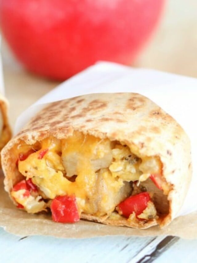 Breakfast burrito cut in half so you can see filling, wrapped in white paper with red apple in background.