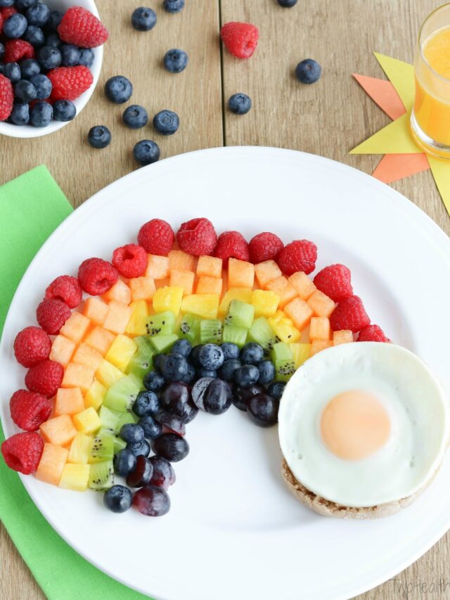 Fruit rainbow with fried egg on white plate with berries, orange juice and green napkin nearby.