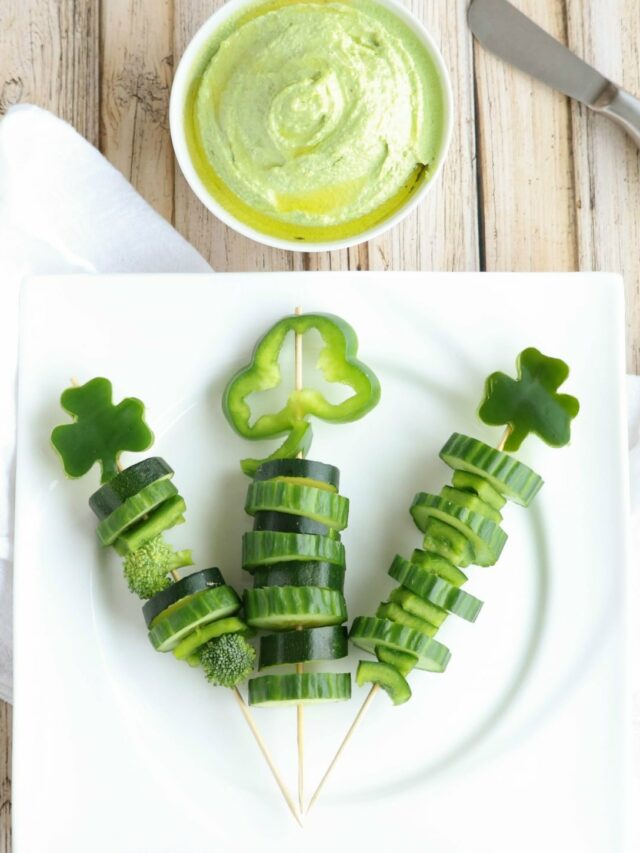 Three skewers with different green veggies on white plate with green dip and spreader nearby.