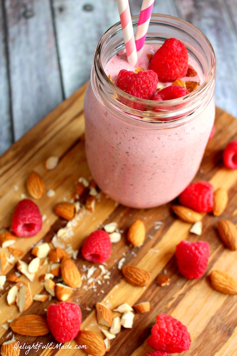 Pink smoothie in mason jar garnished with raspberries, almonds and pink-striped straws with more nuts and berries scattered nearby.