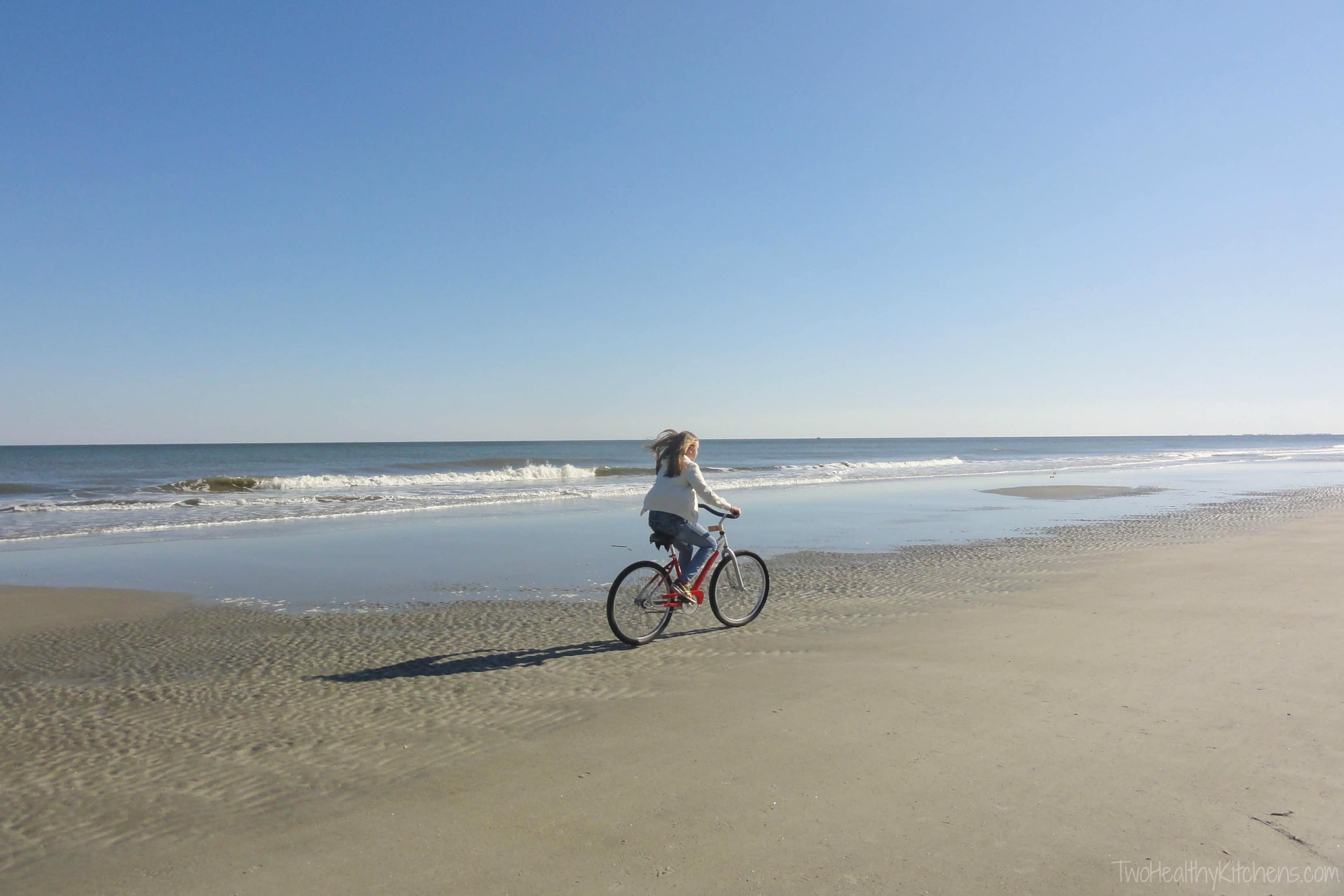 Girl in jeans and jacket riding red bike on beach at surf line.