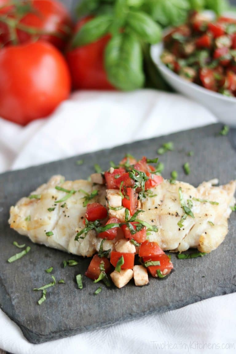 Closeup of one portion of grilled fish with Caprese Topping draped overtop on slate plate with extra tomatoes and basil behind.