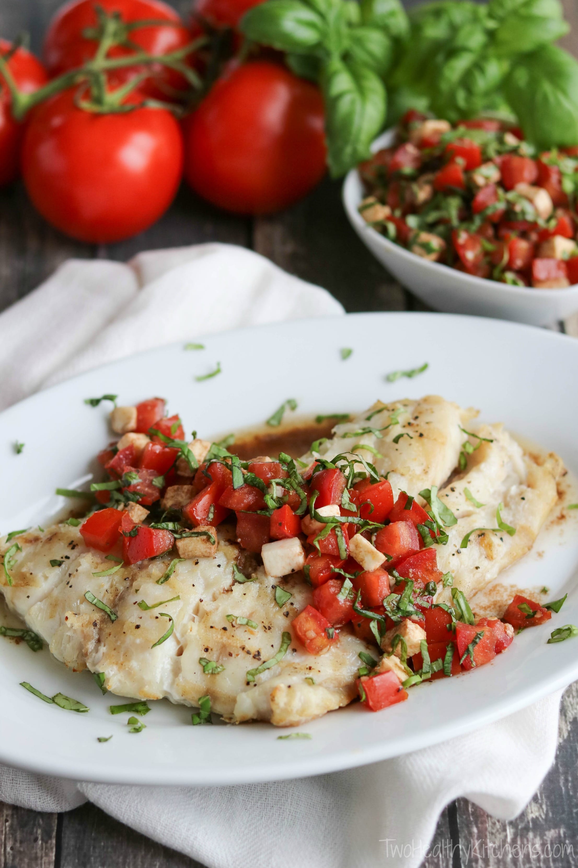 Closeup of whole grilled fish filet on white platter with topping across; small bowl of extra topping and extra tomatoes and basil behind.