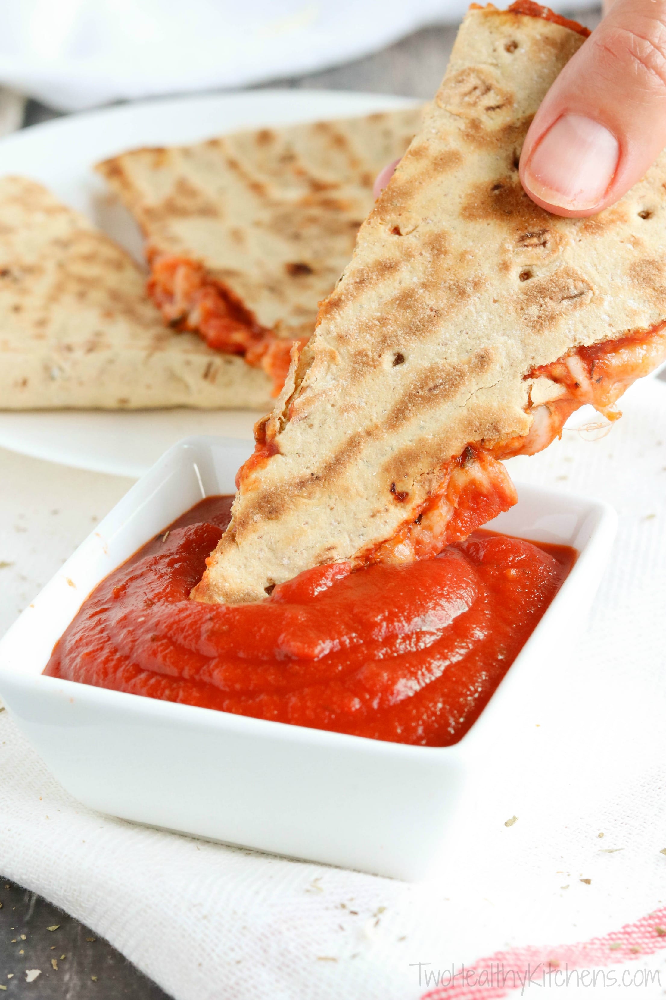 Closeup of hand dipping one quesadilla wedge into little square dish of pizza sauce with more wedges in background.