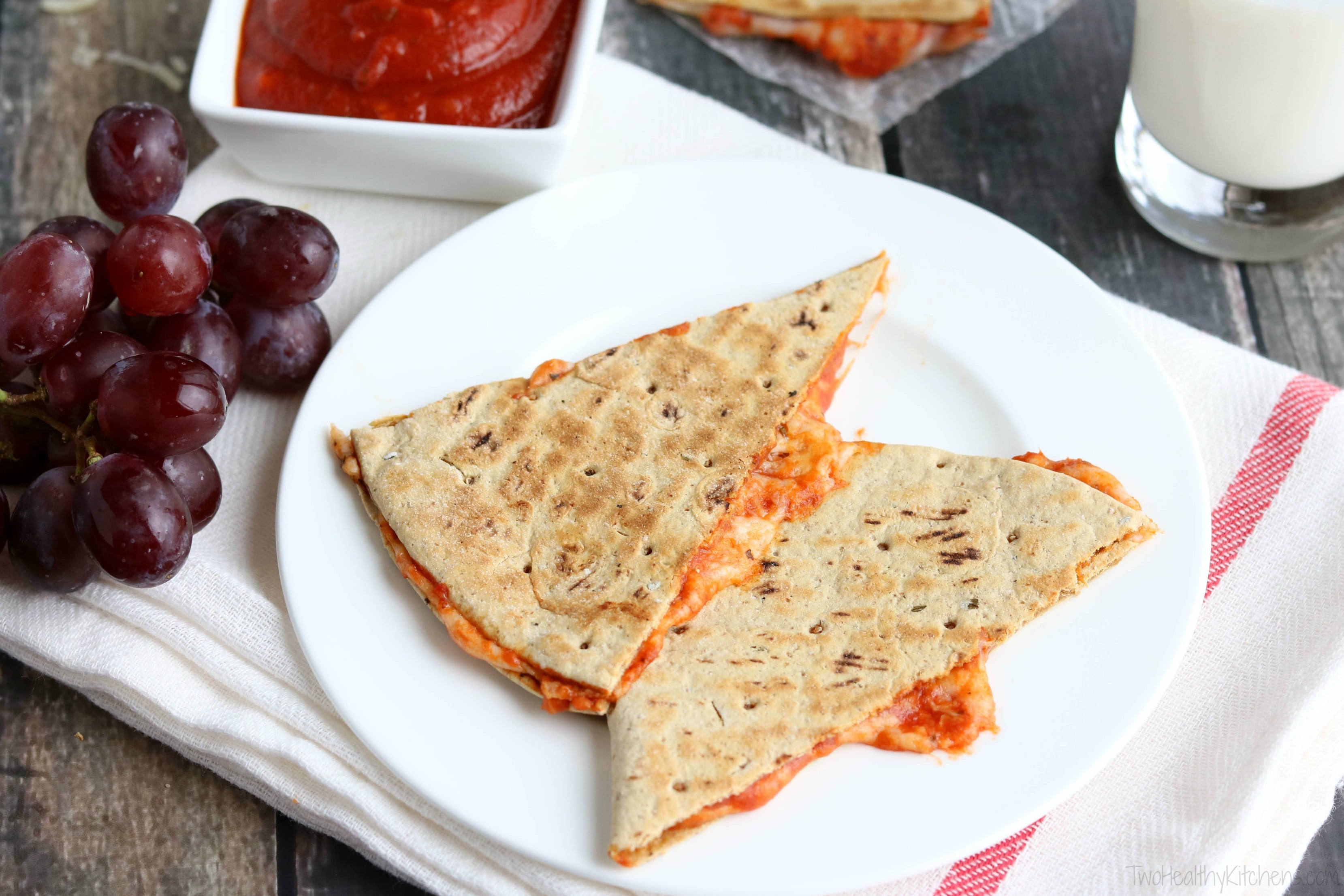 Two quesadilla wedges served on white plate with bunch of grapes, dish of marinara dip and glass of milk surrounding.
